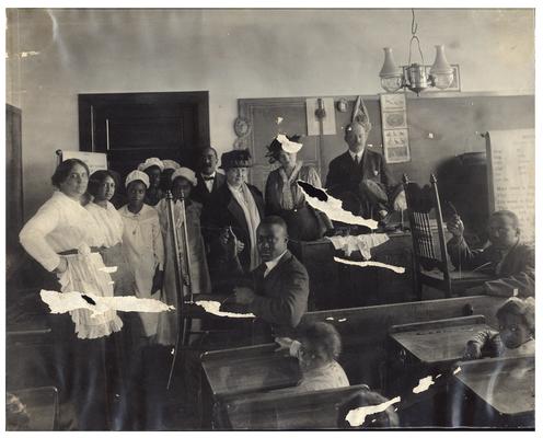 Group portrait of Maddoxtown students at their desks while some demonstrate chair canning. Also present are State School Supervisor F.C. Button, Nannie Faulconer (1865?-1940) and Mattie Dalton. (Two copies)