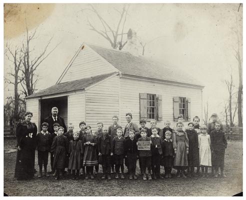 Group portrait of Tyler students in front of the school house with teacher Charlotte Pilcher and M.A. Cassidy, Superintendent of Fayette County Public Schools until 1905. Handwritten on verso, Tyler School-given over to Brech Payne