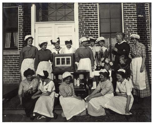 Group portrait of Picadome girls, Nannie Faulconer (1865?-1940) and Belle McCubbing posing with oven and trophy. Handwritten on verso, Nannie Faulconer, Supt.; Dike' Wilkerson 3rd left, standing (sailor collar). Mrs. Belle McCubbing teacher, Willie Carr far right sitting. (Two copies)