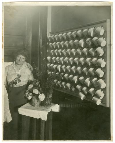 Portrait of Nannie Faulconer (1865?-1940) standing beside a wall of soup cups