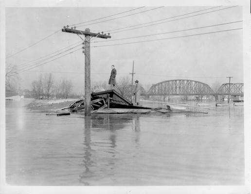 Two unidentified men standing on roof of destroyed building surrounded by flood waters. Railroad bridges visible in the background