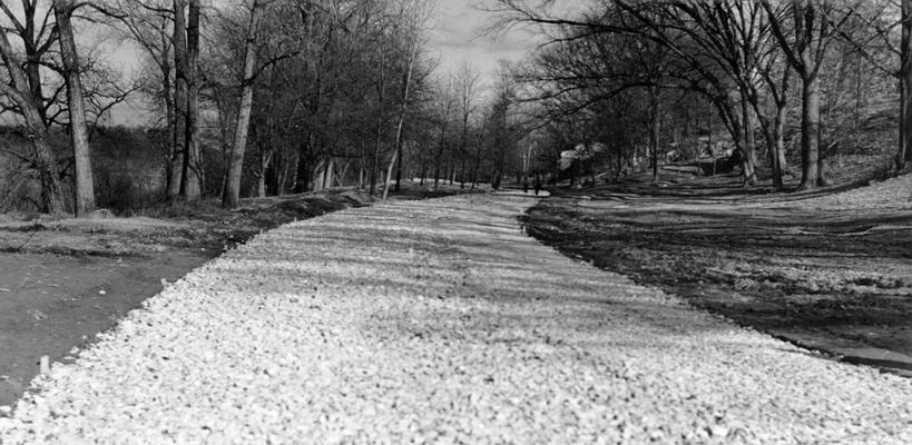 Project #897 District 6: Construction of a cement-bound resurface on the Lower River Road in Shawnee Park, Louisville, KY. The view, photographed January 13, 1936, shows a portion of the road after crushed rock had been placed on it. View is facing north from the foot of Broadway. Shawnee Park is on the right and the Ohio River is on the left