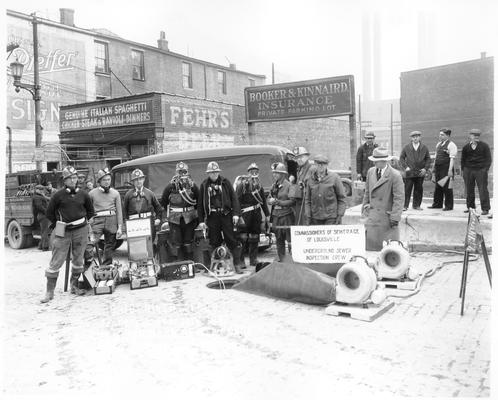 Team of men with helmets, oxygen masks, etc., standing around a manhole with sign reading 