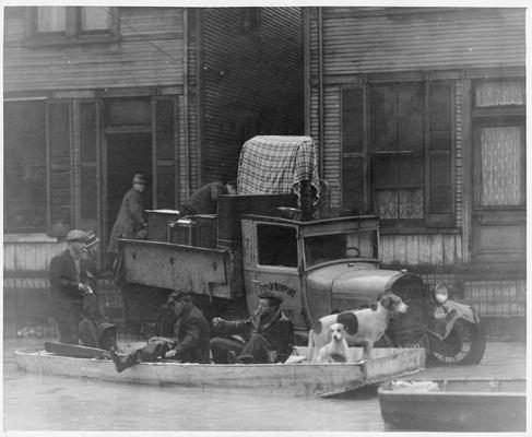 Two men loading belongings onto a truck; two men in a small rowboat with a dog; and two men wading in water - all in front of a flooded building