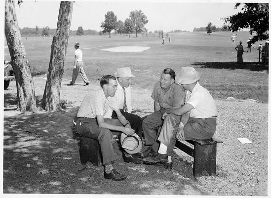 Four men sitting on benches on a golf course. Man in the dark shirt is Ed Kellow, sports editor of the Paducah-Sun Democrat