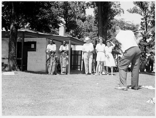 Man is driving a golf ball while several people look on; a refreshment stand is in the background