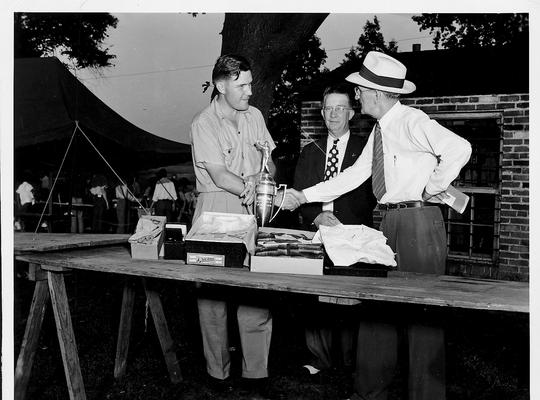 Three men at a table as one receives an award cup. One of the men is Edwin J. Paxton