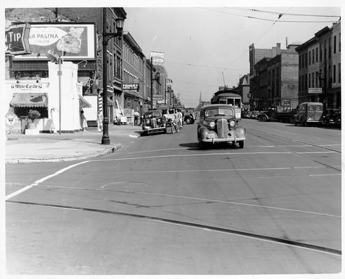 A downtown street with trolley car and several autos.  A White Castle is on the corner