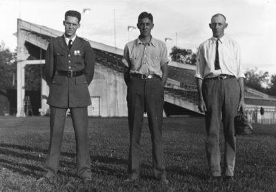 Three men posing in front of stands of McLean Stadium, one military, one medic