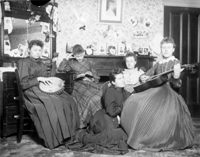 5 women sitting together, 2 are playing instruments while the others listen and 1 is reading