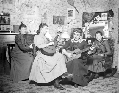 4 women sitting together possibly in a dorm room in Patterson Hall, 2 of them are playing instruments