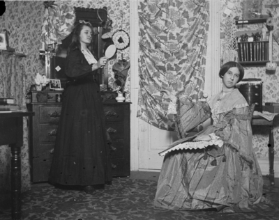 2 women in a room,  one is holding a brush and the other one is sitting in a chair holding a fan and pointing at the other woman