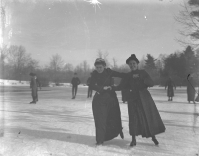 2 women ice skating other people are in the background, possibly at the site of the present student center