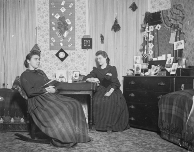 2 women sitting together, 1 is reading a book possibly in a dorm room