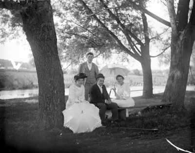 2 women and a man sitting on a bench outside with 1 man standing behind them