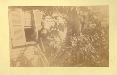 Woman watering plants on the side of a house; Another person looking out window