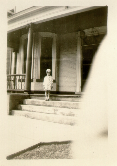Young girl standing on steps of porch wearing hat and coat; (same girl as #127)