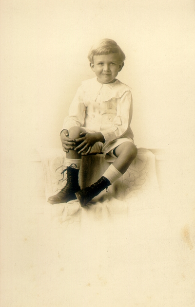 Young boy with grin, wearing white outfit and sitting on bench, printed on post card