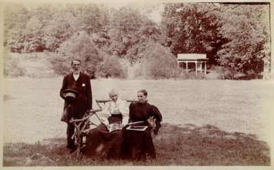 Mrs. M.C. Lyle sitting next to an elderly woman (possibly her mother) with an elderly man standing beside them; Setting in open field with trees and gazebo in background