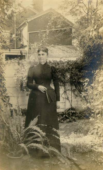 Woman in full black dress standing in garden holding a fan