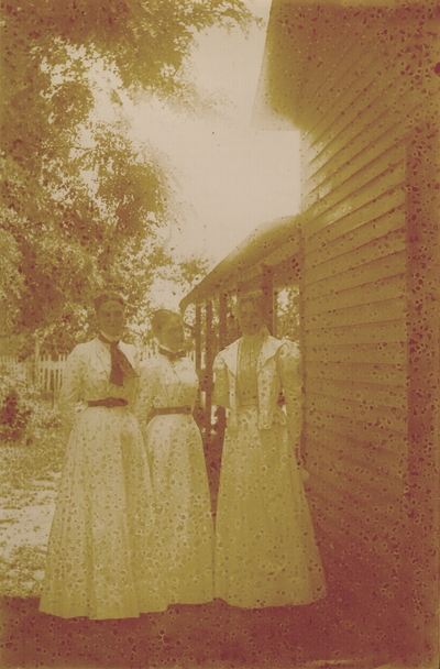 Three young ladies standing beside a house wearing white dresses
