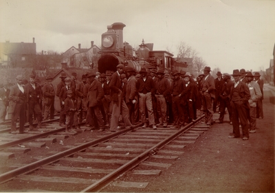 Large group of men standing on or near railroad tracks in front of steam locomotive