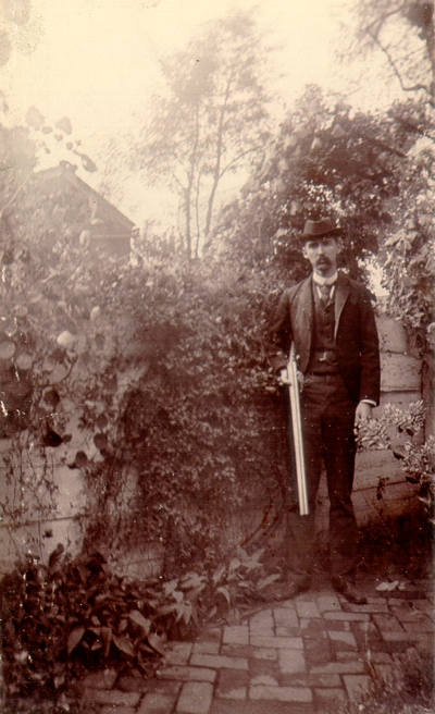 Charles N. Lyle standing in garden in front of fence holding a shotgun