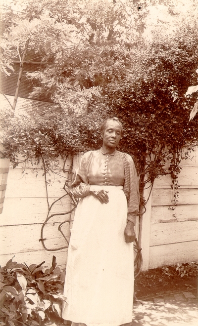 African-American woman with long white apron standing in garden area in front of fence