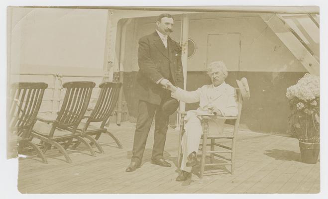 Photograph of Mark Twain seated on a ship, shaking hands with an unidentified man
