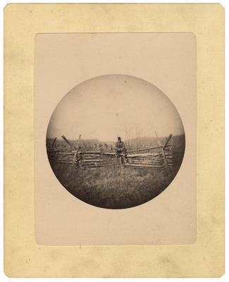 Unidentified man sitting on split-top fence, gun in lap; loose in front of album