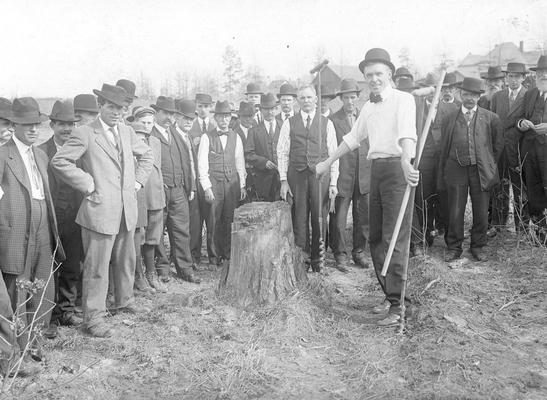 Men standing around a tree stump, one man is holding two augers in his hand, augers are used to create holes in wood and earth