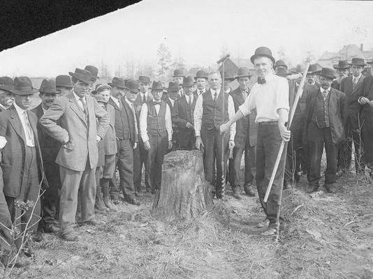 Men standing around a tree stump, one man is holding two augers in his hand, augers are used to create holes in wood and eart