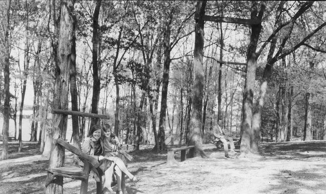 Two young women seated on a park bench adjacent to a man seated on a bench