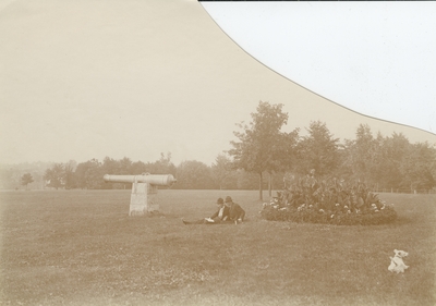 2 unidentified men sitting on the grass near the cannon on the U.K. campus, a small dog is in the photograph as well; corner of photograph is cut away