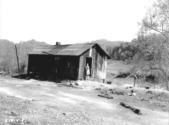Woman standing by a doorway to a shack next, to dirt road, in the country