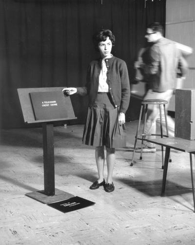 Woman standing next to TV billboards being used for still shots for introduction of Survey of English Literature televised course. (Kathy Roper White)