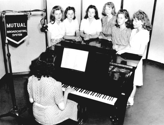 A group of six women gathered around a studio piano singing: Mutual Broadcasting System banner attached to microphone stand
