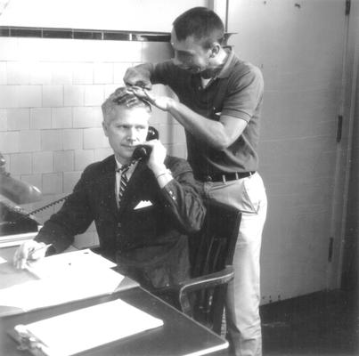 Man pretending to be cutting the hair of man sitting at desk (Jerry Kuykendall, Stu Hallock)