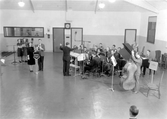 UK Radio Studios: Band being conducted while two sets of people stand at microphones (Mutual Broadcasting system banner hanging on first microphone)