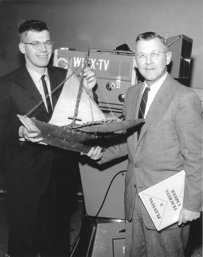 Student and teacher posed in front of WLEX-TV studio camera holding model of wooden ship, John P. Malick (Dr. Ellis Hartford)