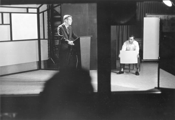 Cultural anthropology professor Dr. Doug Schwartz at lectern in television studio; man sitting right possibly holding cue-card or billboard (photo taken from control booth)