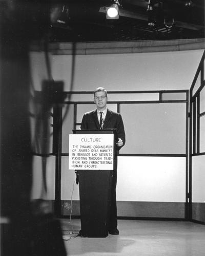 Dr. Doug Schwartz  speaking at lectern in front of camera; card on lectern has definition of culture