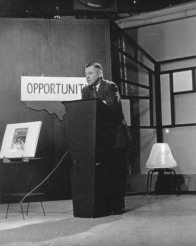 R. Ellis Hartford standing at lectern in television studio; sign behind him reads 