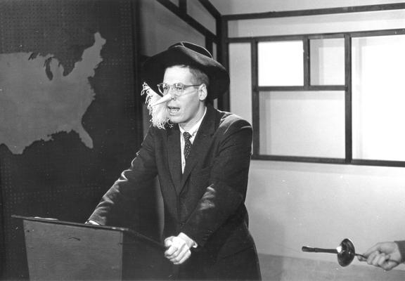 Dr. Doug Schwartz at lectern with nose and hat, as Cyrano