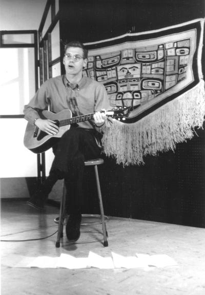 Dr. Doug Schwartz playing guitar in front of Pre-Columbian tapestry, with music lyrics on floor in front of him