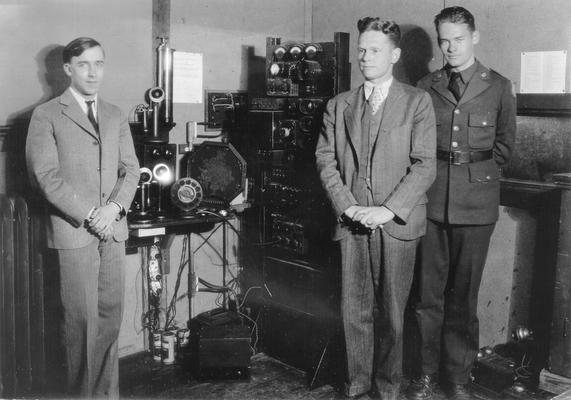 Three men standing, one in uniform, around telephone and radio equipment (microphone on table next to telephones is American Carbon microphone)