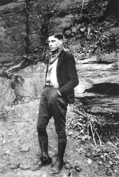 Man wearing boots, jacket, tie, and handkerchief with pistol holstered behind belt; standing near rock wall