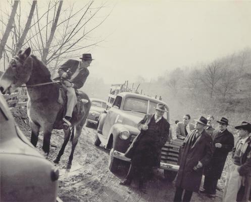 Group of men standing around trucks on dirt road, talking to man on mule holding, flask of whiskey