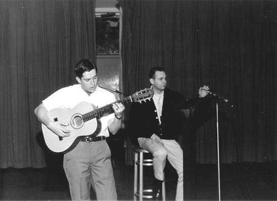 Young man playing guitar while another man adjusts microphone