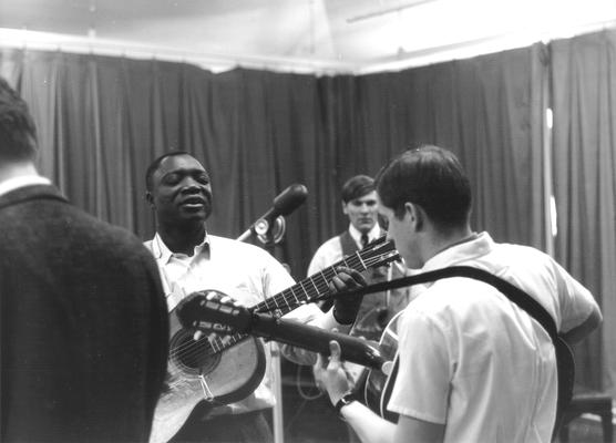 African-American man playing guitar and singing; second guitarist and spectator in background